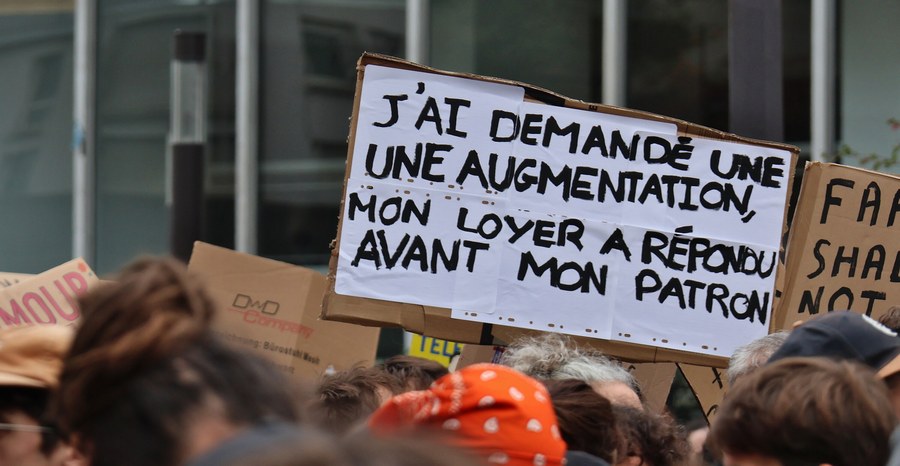 A sign above the march in Paris in October 2026 reads "I asked for a raise, my rent answered before my boss."